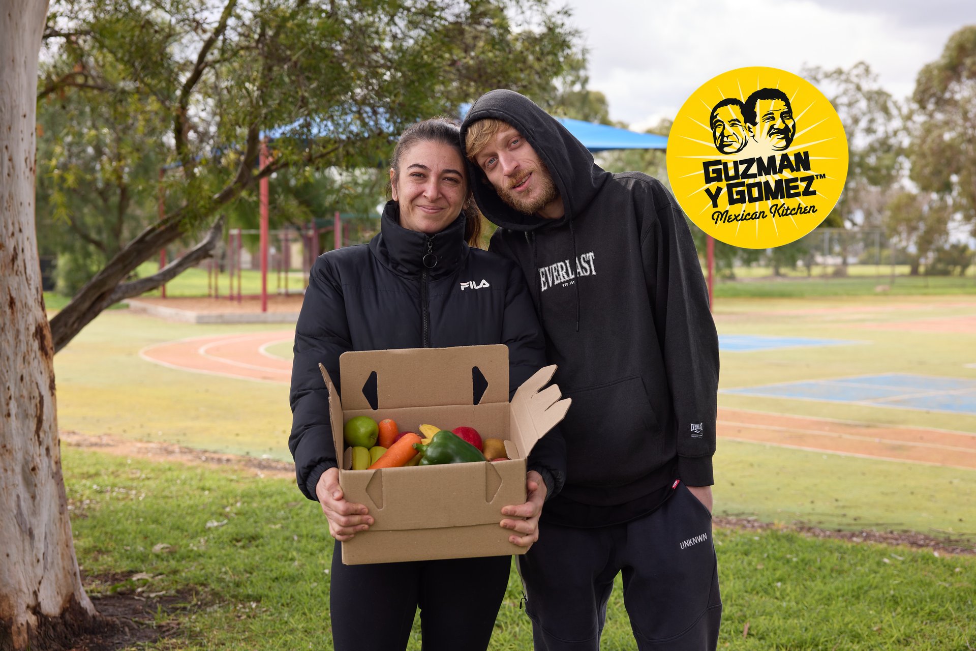 A couple holding a The One Box fresh food box, with the Guzman y Gomez logo